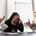 Frustrated millennial female worker sitting at table with colleagues, felling tired of working quarreling at business meeting. Upset stressed young businesswoman suffering from head ache at office.