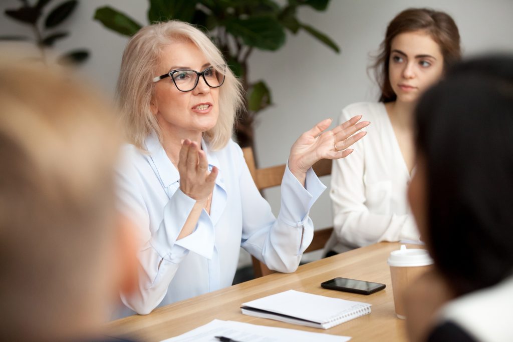 Attractive aged businesswoman, teacher or mentor coach speaking to young people, senior woman in glasses teaching audience at training seminar, female business leader speaker talking at meeting