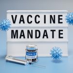 The words "vaccine mandate" displayed on a lightbox. Syringe, virus models and a bottle of vaccine around.