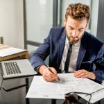 Handsome tax manager dressed in the suit working with documents and laptop at the modern office interior