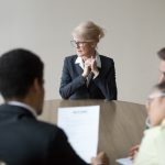 Middle aged stressed woman sitting at the desk in office at meeting. Young multiracial workers team sitting their backs to camera. Passing interview, hiring, recruiting and age discrimination concept