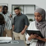 Young Muslim businesswoman in hijab holding black folder with documents against group of intercultural colleagues mocking at her